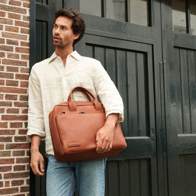 PLEVIER Mann in lässiger Kleidung hält eine braune Tasche, steht vor einer Ziegelmauer.